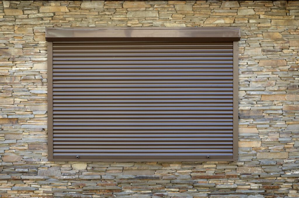 A window with brown metal rolling shutters. Wall decoration with artificial flatten stone